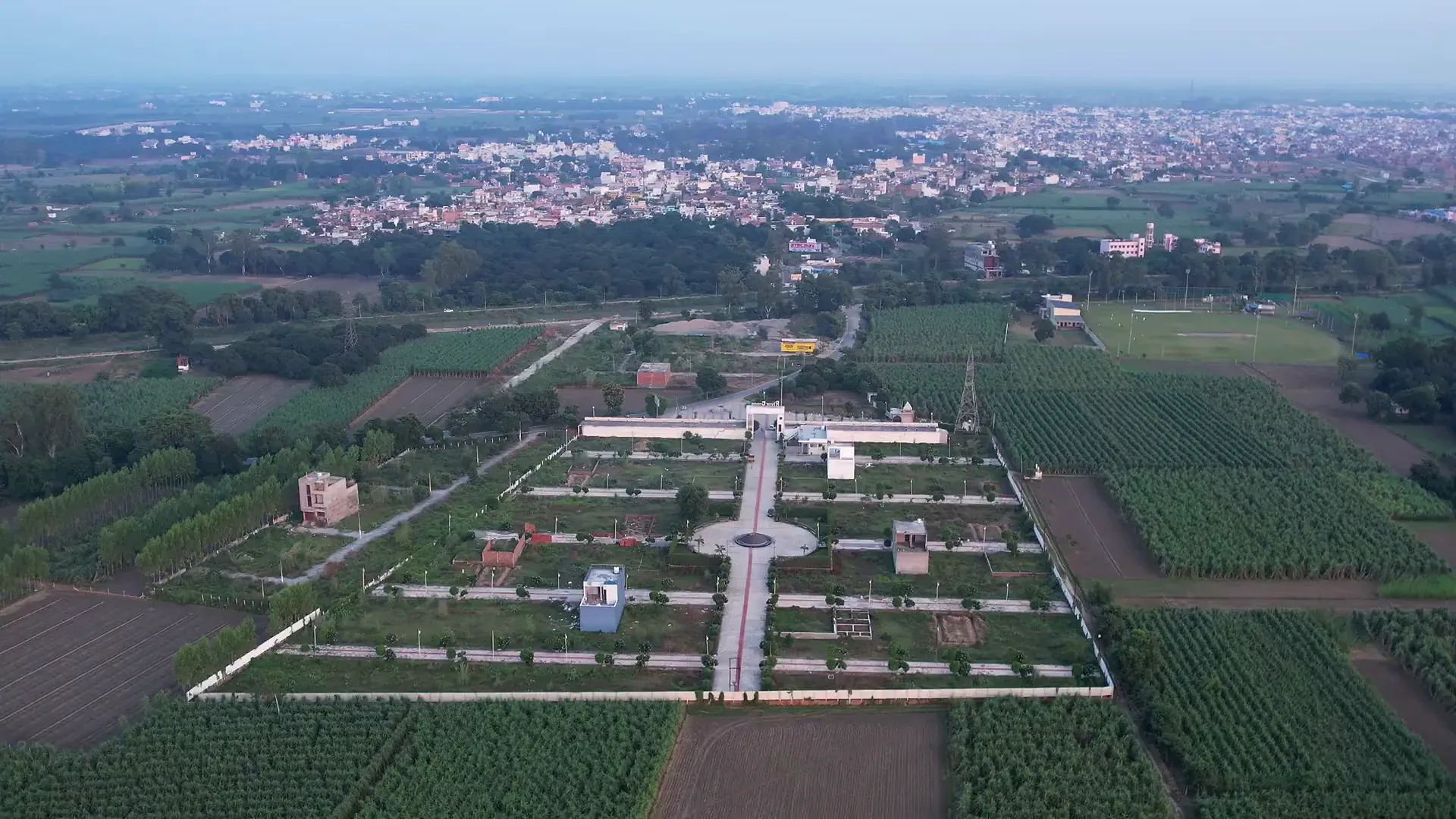 Aerial view of River Green Society: the walled grid of plotted streets, the fountain roundabout and the entrance, set among green fields with the town beyond