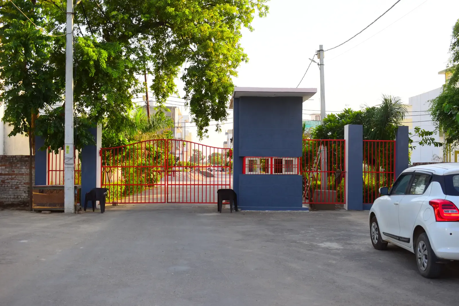 Gayatri Enclave entrance with red gates and a guard cabin, a car parked outside