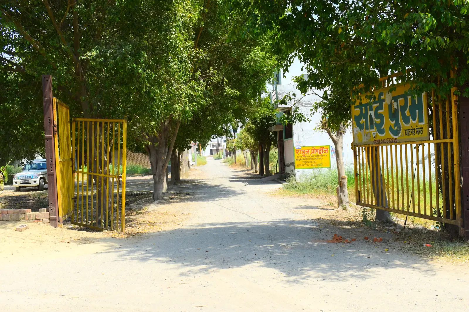 Saipuram: tree-shaded entrance lane with open yellow gates and the colony signboard