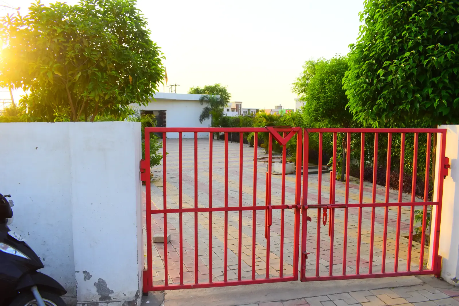 Gayatri Enclave: red metal gate in a white boundary wall, sunlight flaring behind