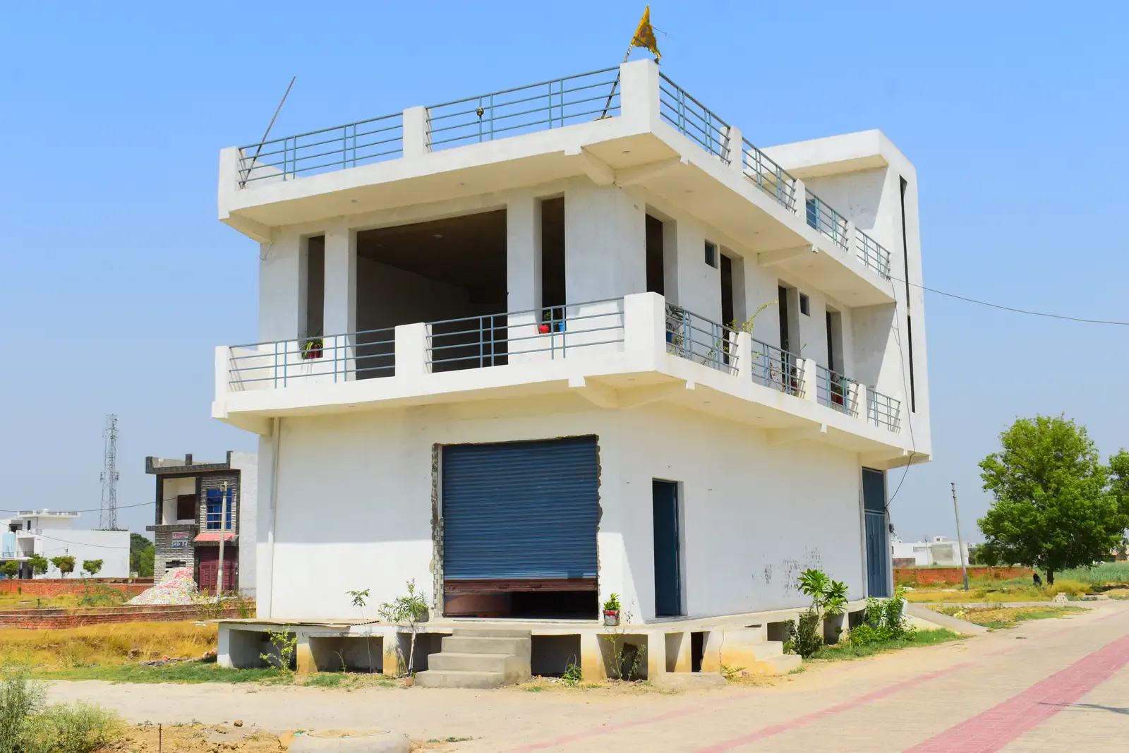 Basant Kunj: two-storey white corner building with balconies and a blue rolling shutter
