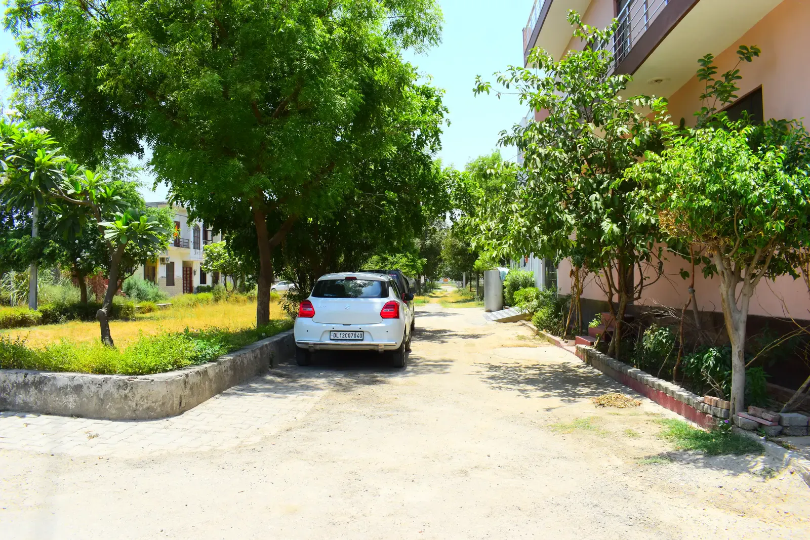 Defence Garden Meerut: shaded internal street with mature trees, a lawn verge and a parked car