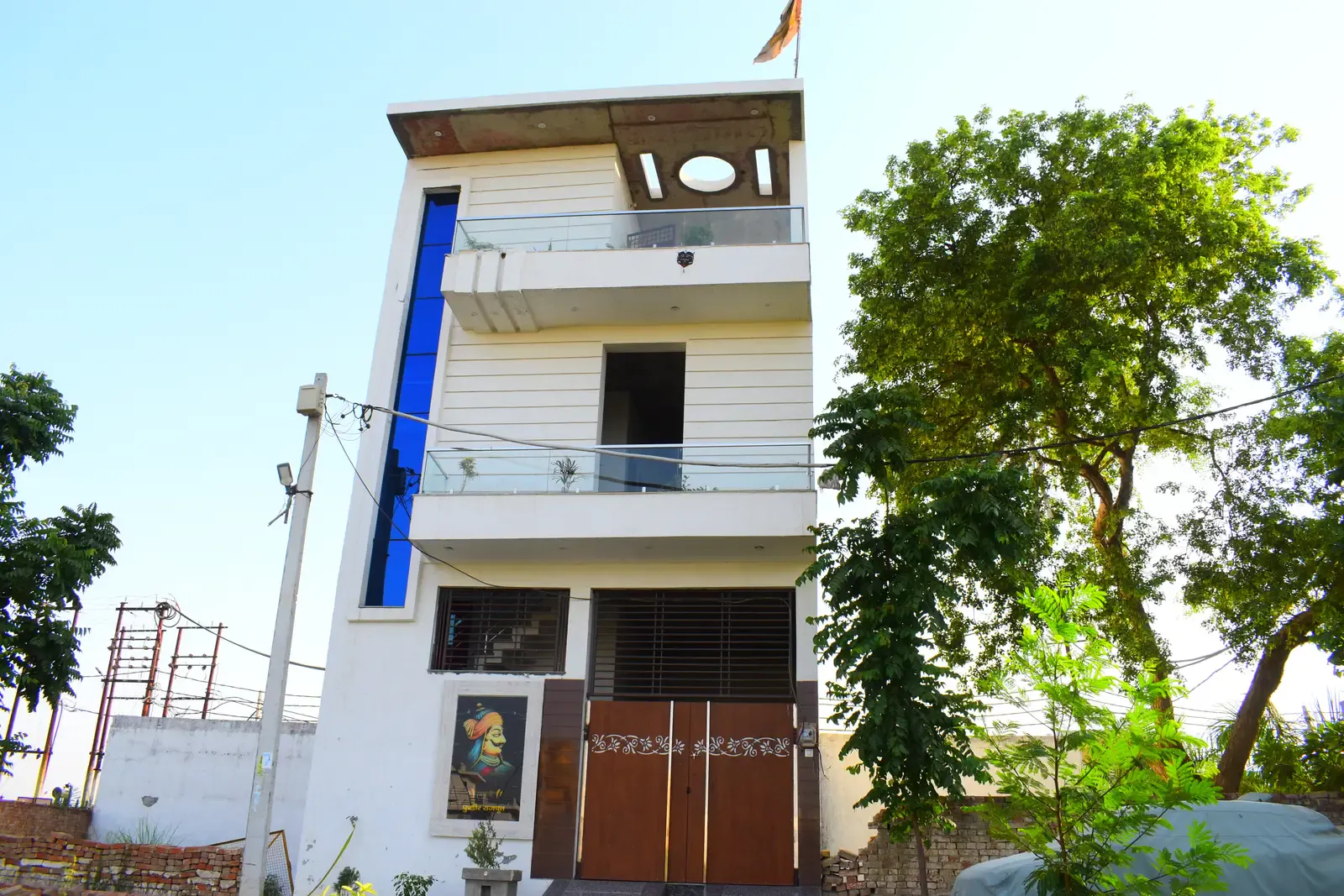 Gayatri Enclave: narrow three-storey white home with a blue glass strip beside a large tree